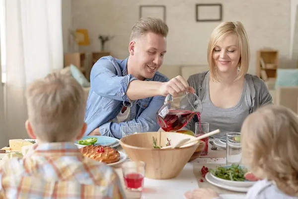 Blond Caucasian man pouring homemade fruit drink into glass while sitting at table and having dinner with his beautiful wife and children at home