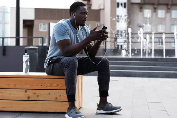 Aktif kıyafetli genç Afrikalı sporcu şehir merkezinde antrenmandan sonra dinleniyor, bankta oturuyor, akıllı telefondan mesaj yazıyor ve müzik dinliyor.
