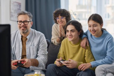 Dört kişilik aile oturma odasında kanepede oturup video oyunu oynuyorlar.