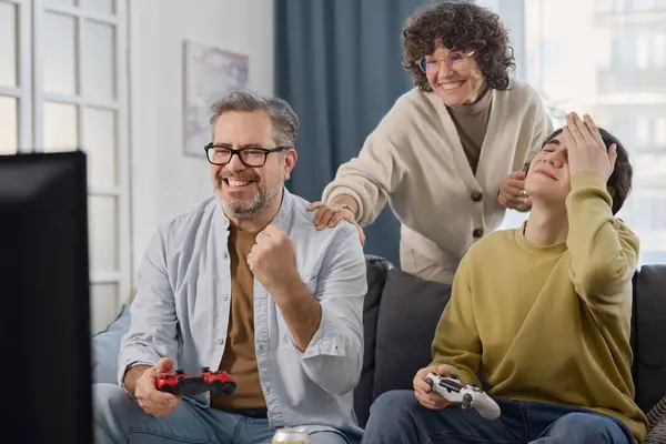 Happy dad playing console game with his son while mother supporting them while they sitting on sofa at home