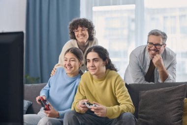 Mutlu çocuklar video oyunu oynamak için joystickleri kullanırken ebeveynler arkalarında duruyorlar.