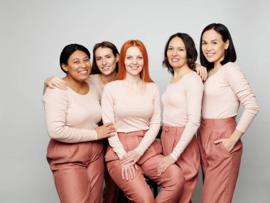 Portrait of smiling interracial women in same pink outfits standing together against isolated background clipart