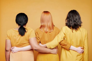 Sarı arka planda, farklı renkte saçları olan farklı kadınların birbirini kucaklaması.