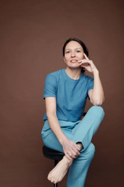 Mavi elbiseli, gülümseyen orta yaşlı bir kadının portresi taburede oturuyor ve koyu arkaplana dokunuyor.