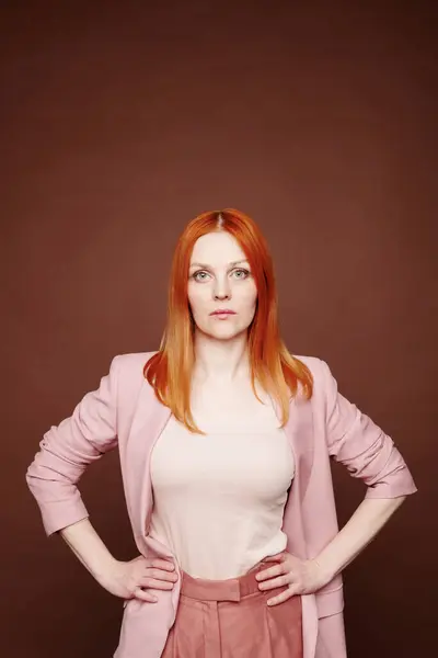 Portrait of serious young redhead woman in pink jacket holding hands on hips against brown background