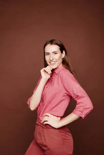 Portrait of cheerful confident young lady in pink shirt touching chin against brown background