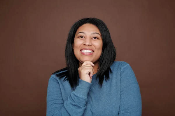 Portrait of excited sincere young black woman laughing while touching chin against brown background