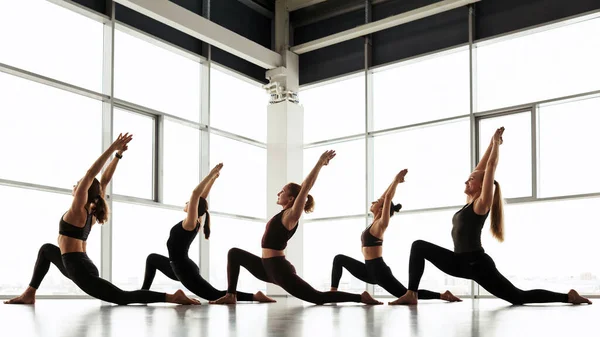 Group of young women in black sportswear doing crescent low lunge pose against panoramic window in studio
