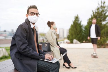 Gözlüklü ve maskeli yakışıklı Arap bir adamın portresi. Şehir parkında bankta oturuyor ve dizüstü bilgisayarında yazı yazıyor.