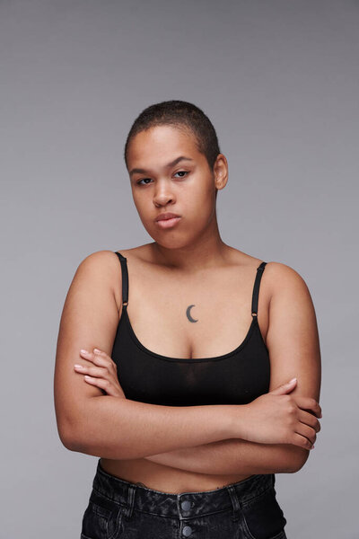 Portrait of serious body positive black woman with tattoo standing with crossed arms against gray background