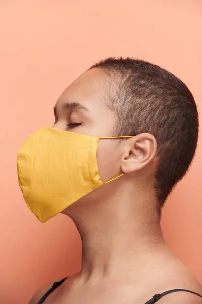 Side view of depressed young black woman with buzz cut wearing yellow mask and keeping eyes closed against orange background, coronavirus concept