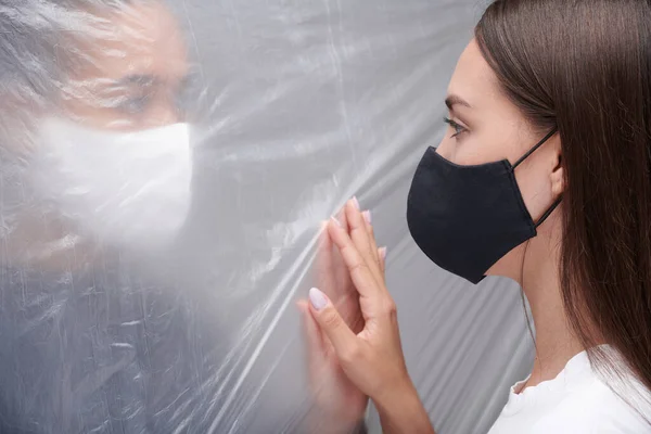Young multi-ethnic women in cloth masks touching through polyethylene wall while having barrier during coronavirus epidemic