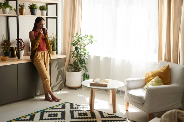 Stock image Calm young woman in casual outfit leaning on counter and drinking tea in living room