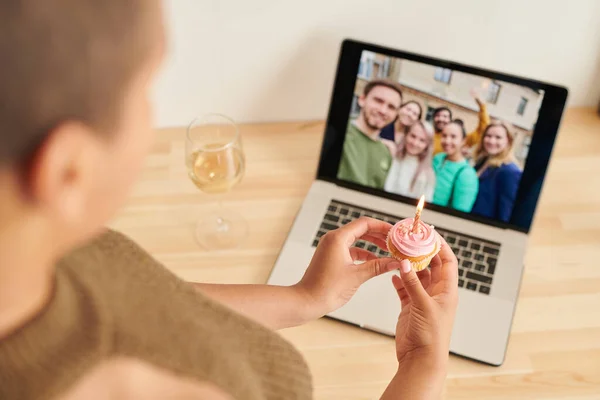 Over shoulder view of black woman holding cupcake with burning candle and celebrating birthday with friends via video link