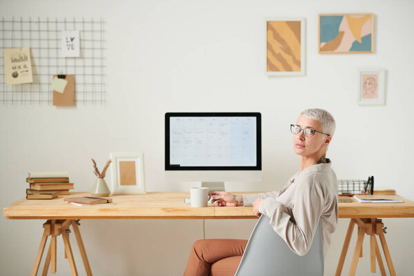 Portrait of content middle-aged businesswoman with short blond hair sitting at desk with computer and drinking coffee
