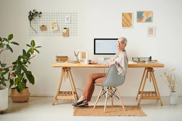 Confident middle-aged lady in eyeglasses sitting at desk with modern computer in cozy office