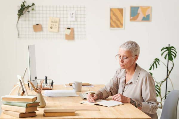 Busy middle-aged businesswoman in eyeglasses sitting at desk and making notes in planner while working in modern office