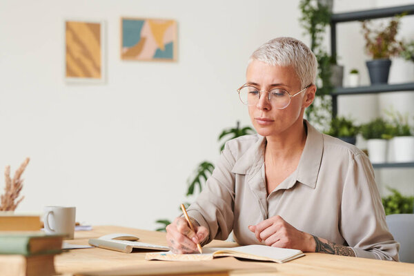 Serious short-haired lady in glasses sitting at wooden desk in cozy home office and making notes in diary while planning day