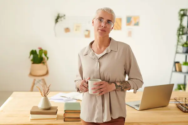 Portrait of content hipster middle-aged lady in round glasses holding mug of coffee in modern office