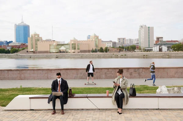 Modern business people in facial masks sitting on bench on embankment and working with devices outside