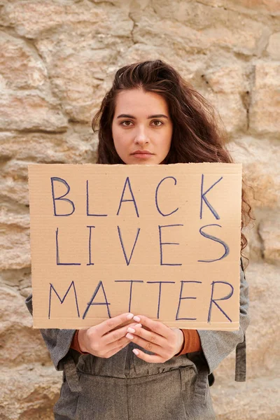 Portrait of serious young woman standing with banner against stone wall and appealing to respect black people