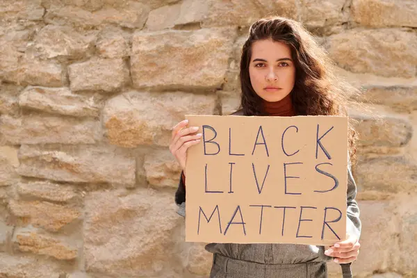 Portrait of serious young woman showing Black lives matter banner against stone wall