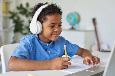 Kablosuz kulaklıklı içerikli siyah çocuk masada oturuyor ve özel öğretmenle çalışırken dizüstü bilgisayar kullanıyor.