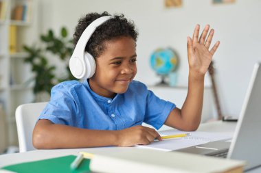 Mavi tişörtlü sevimli Afro-Amerikalı çocuk. Masada oturuyor ve online derste hocayı selamlarken el pençe divan duruyor.