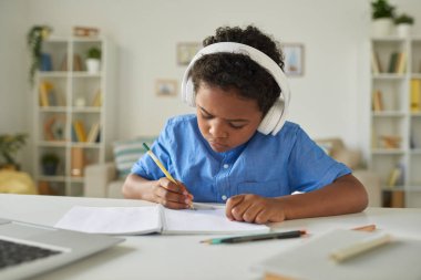 Serious African-American schoolboy in headphones sitting at desk and making notes while listening to tutor online clipart