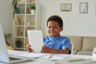 Hoş Afro-Amerikalı çocuk oturma odasında oturmuş video konferans uygulaması ile iletişim kurarken tablet kullanıyor.