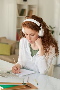 Kablosuz kulaklıklı, odaklanmış kızıl saçlı kız masada oturmuş, online dersler dinlerken notlar alıyor.