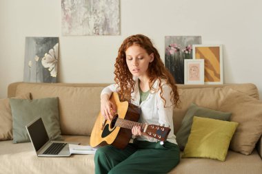 Kanepede oturup gitar akorlarını öğrenirken dizüstü bilgisayar kullanan kızıl saçlı kız.