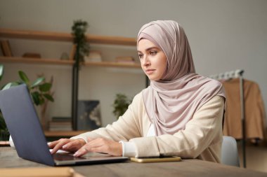 Başörtüsü takan çekici Orta Doğulu kadının yatay portresi modern ofis odasında dizüstü bilgisayarla çalışıyor.