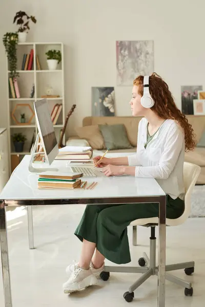 Kablosuz kulaklıklı, ciddi kıvırcık saçlı kız masada oturuyor ve oturma odasında iş yaparken internetten özel ders dinliyor.