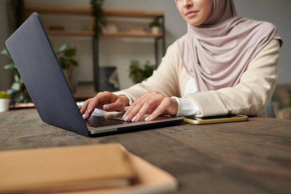 Modern Muslim woman wearing hijab sitting at office desk typing something on her laptop, copy space