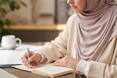 Masada oturan soluk pembe başörtüsü takan Müslüman kadın defterine not alıyor.