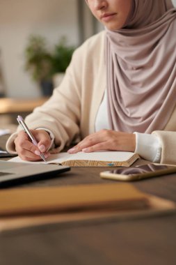 Orta Doğulu kadın başörtüsü takıyor ve abaya ofis masasında oturmuş not defterine notlar yazıyor, dikey kafasından vurulmuş.