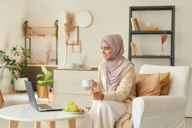 Modern Müslüman kadın evde çay içerek ve dizüstü bilgisayardan komedi filmi izleyerek vakit geçiriyor.
