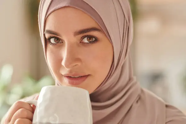 Attractive Muslim woman wearing pale pink hijab on head holding cup of coffee looking at camera, horizontal close-up portrait