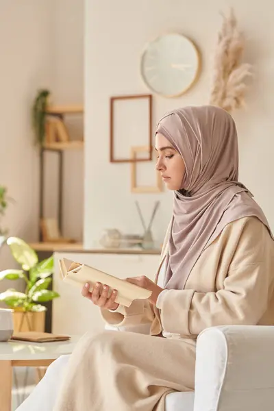 Modern Muslim woman wearing pale pink hijab and casual clothing sitting in armchair reading interesting book