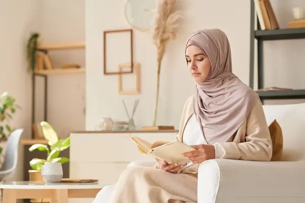 Beautiful Muslim woman wearing pale pink hijab and casual clothing sitting relaxed in armchair reading book, copy space