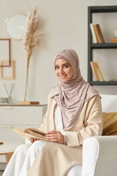 Vertical portrait of joyful young Middle Eastern woman wearing hijab sitting in armchair holding book smiling at camera