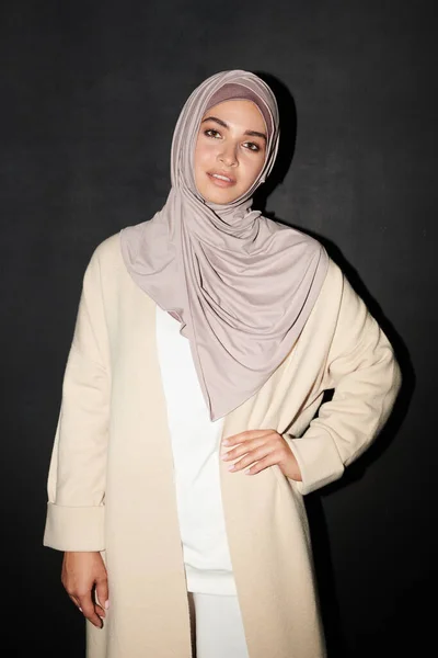 Vertical medium long portrait shot of confident Muslim girl wearing hijab standing against black wall background with hand on hip