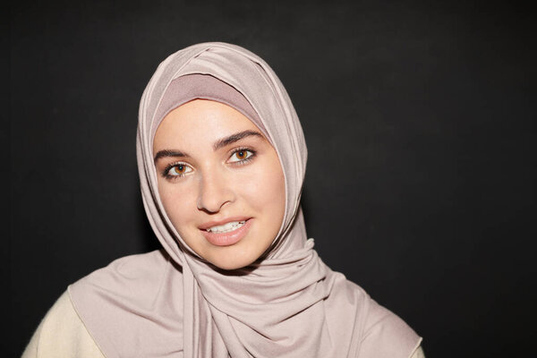 Head and shoulders portrait of beautiful Muslim girl wearing hijab standing against black wall background smiling at camera