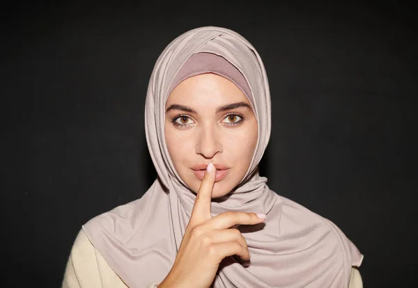 Horizontal head and shoulders shot of modern Muslim woman wearing hijab with fore finger over lips making silence gesture