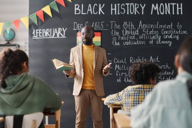 Afro-Amerikan öğretmen tahtaya karşı duruyor ve sınıftaki öğrencilere Siyahi Tarih Ayı 'ndan bahsediyor.