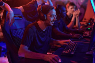 Şapkalı ve gözlüklü gülümseyen genç profesyonel oyuncu Esport turnuvasına katılırken bilgisayar kullanıyor.