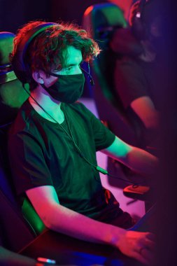 Maskeli ve kulaklıklı, karanlık odada oturmuş video oyunu oynayan odaklanmış beyaz bir adam.