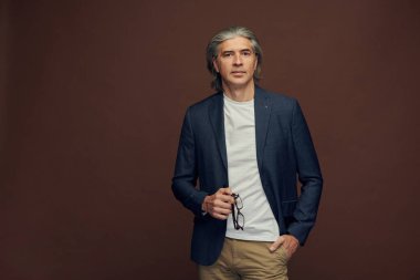 Kahverengi arka planda duran, zarif gözlüklü, güler yüzlü, gri saçlı adamın portresi.