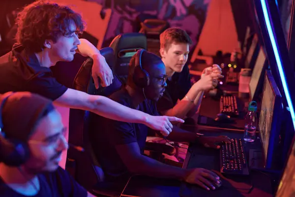 Young man pointing at computer screen while giving advice to African-American gamer focused on game in esports club